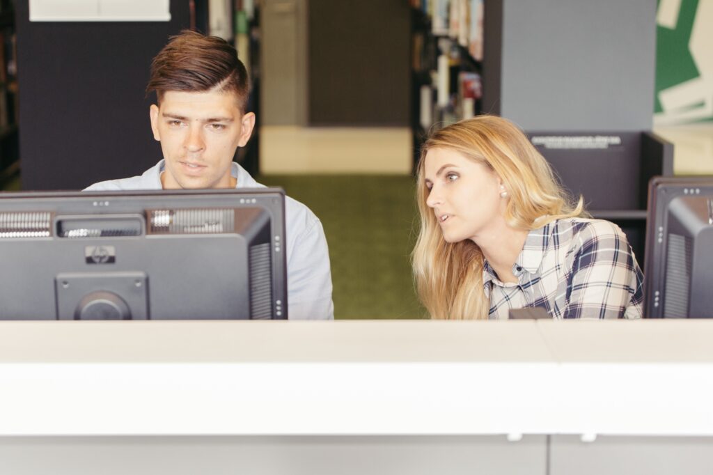 Two employees in front of their computers while sitting side-by-side talking about informing colleagues of the office move in the concept of 'How to Inform Employees About an Office Move'.